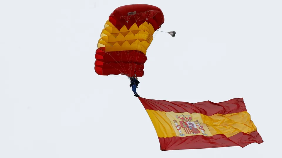 Paracaidista suspendido de un paracaídas rojo y amarillo que transporta una bandera de España desplegada.