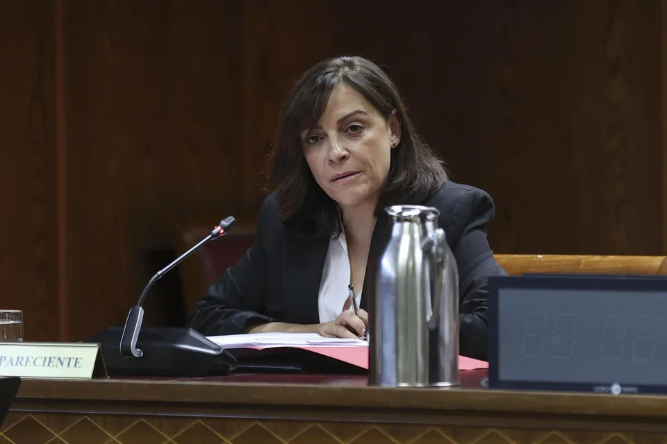 Mujer con chaqueta oscura sentada ante un micrófono durante una comparecencia, con documentos sobre la mesa y una jarra metálica en primer plano.