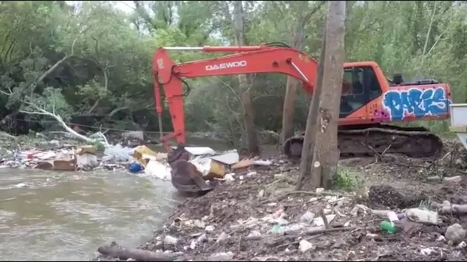 Una excavadora roja trabaja junto a un cauce de agua rodeado de residuos y vegetación.