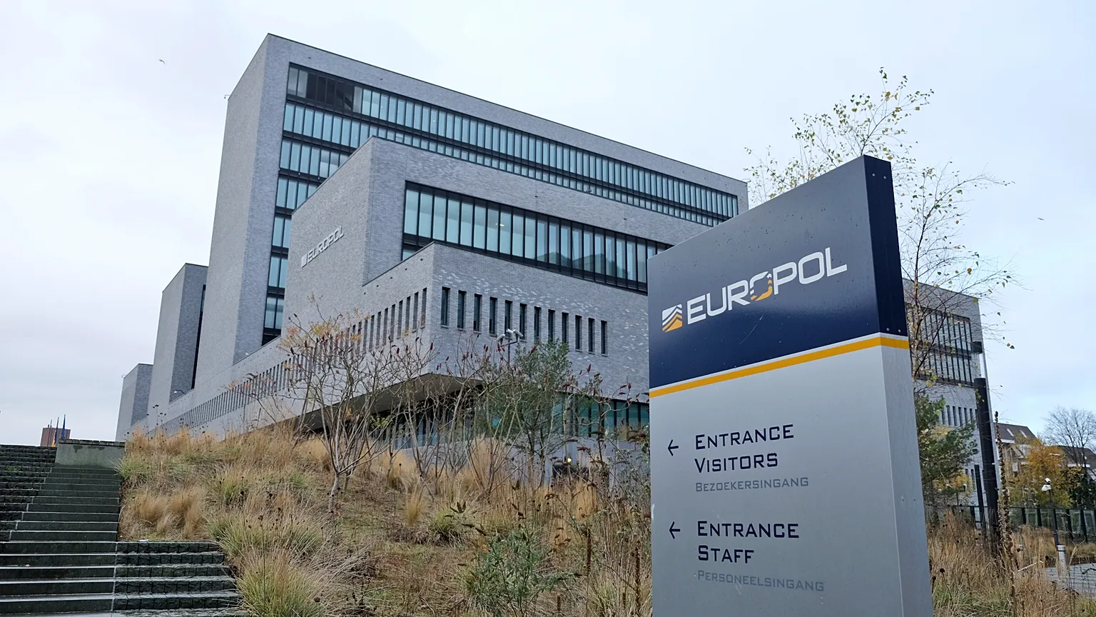 Vista exterior de la sede de Europol en La Haya, con la señal EUROPOL y los accesos indicados para visitantes y personal.